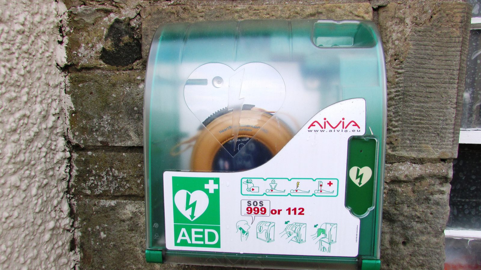 A green public-access AED cabinet mounted on a stone wall